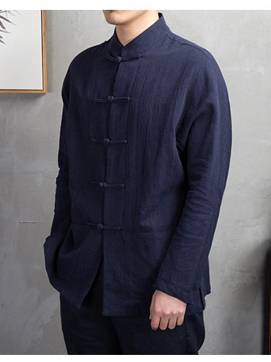 A man wearing a dark navy blue, long-sleeve, mandarin-collar tang kongfu shirt with traditional Chinese frog closures, shown from the neck down against a textured gray wall.