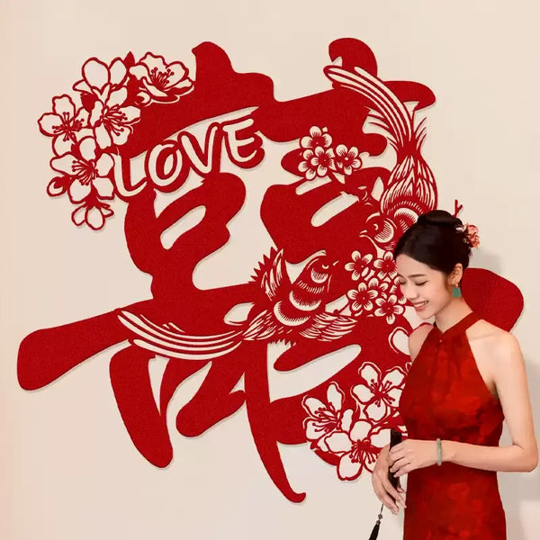 Woman in a red dress standing next to large red Chinese wedding double happiness wall decor  Features a pair of birds, blooming flowers, and the word “LOVE” within the classic Double Happiness symbol – representing harmony, romance, and joyful union, against a beige background.