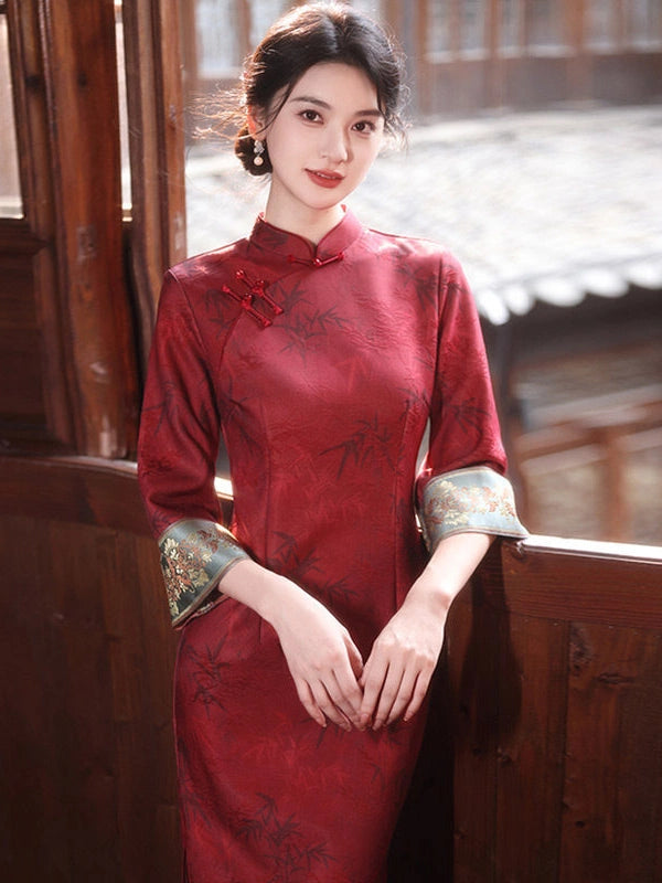 A woman wearing a red embroidered cheongsam-style dress with patterned green and gold cuff accents, standing in a traditional wooden courtyard setting.