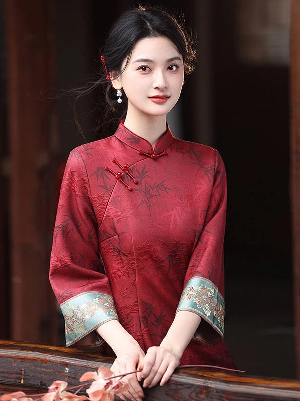 A woman wearing a red Chinese cheongsam jacket with tonal floral embroidery and ornate light blue patterned cuffs, posed in front of a dark wooden background.