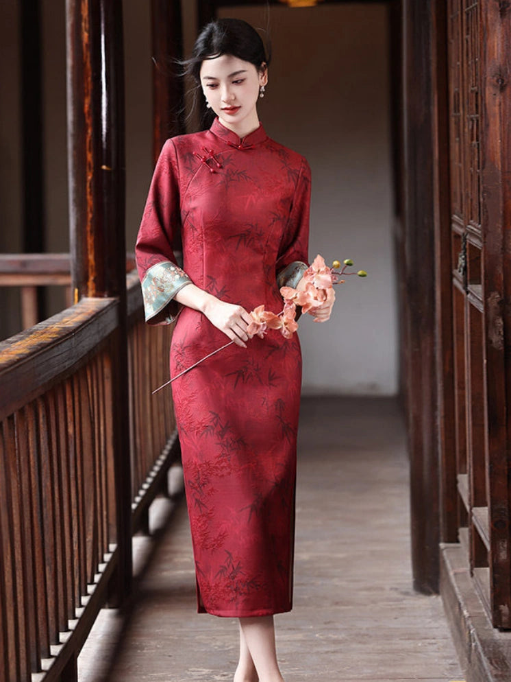 A woman wearing a red Chinese cheongsam dress with black floral patterns and green patterned three-quarter sleeves, standing in a traditional wooden corridor and holding a small bouquet of flowers.