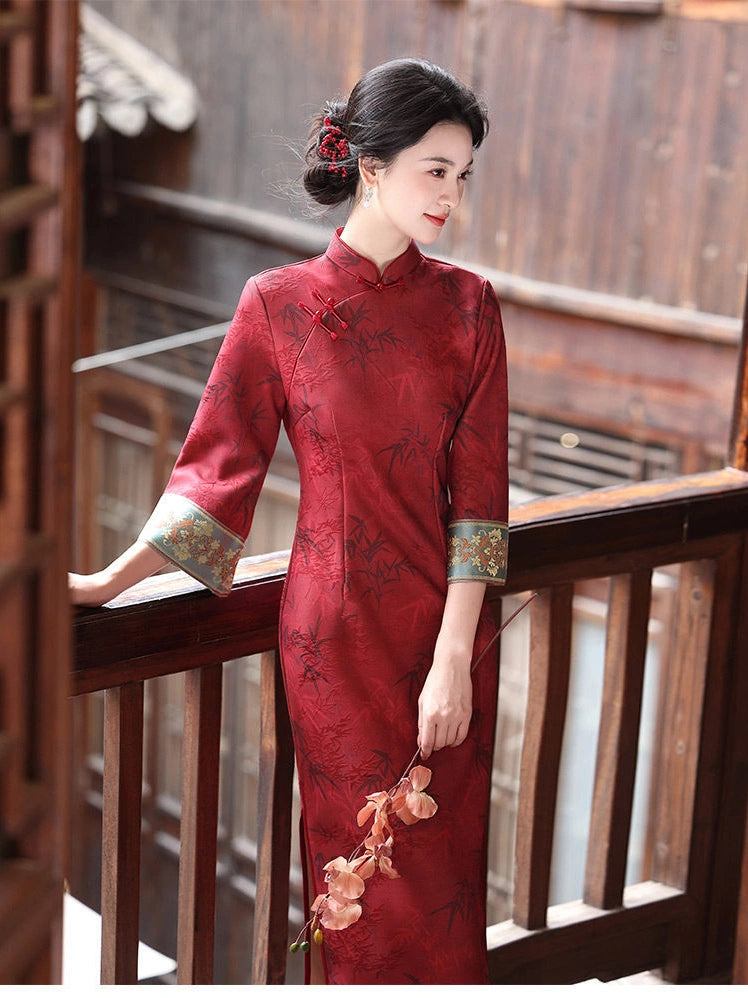 A woman wearing a red embroidered cheongsam/qipao dress with green patterned cuffs, standing on a wooden balcony.