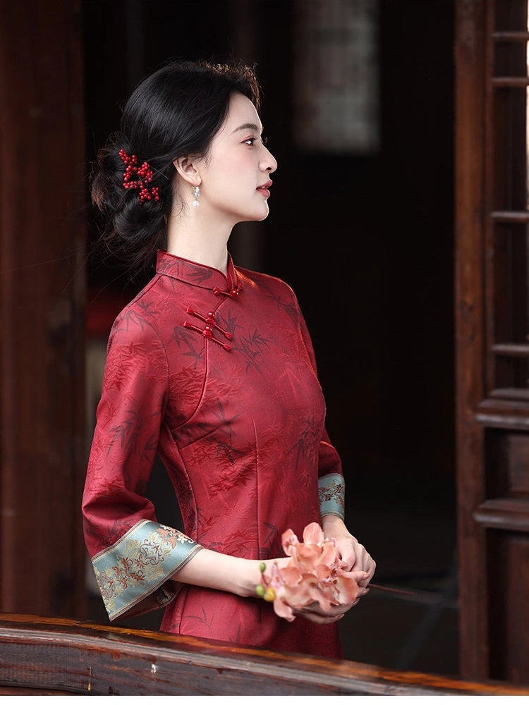 A woman in profile wearing a Crimson Red Bamboo Jacquard Long Sleeve Qipao With Embroidered Cuff , standing in a dark wooden doorway and holding a pink flower bouquet.