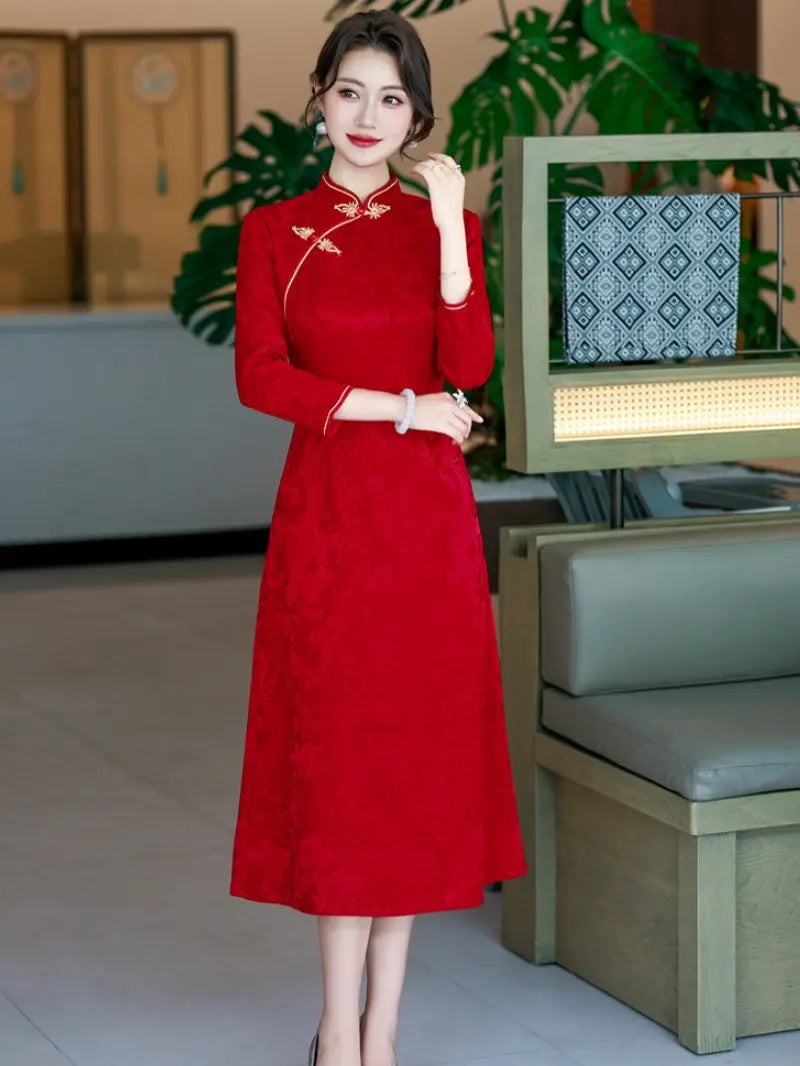 A woman wearing a red long-sleeve a line qipao cheongsam-style dress with gold embroidery and a high collar, standing in an indoor modern setting with plants and furniture visible.