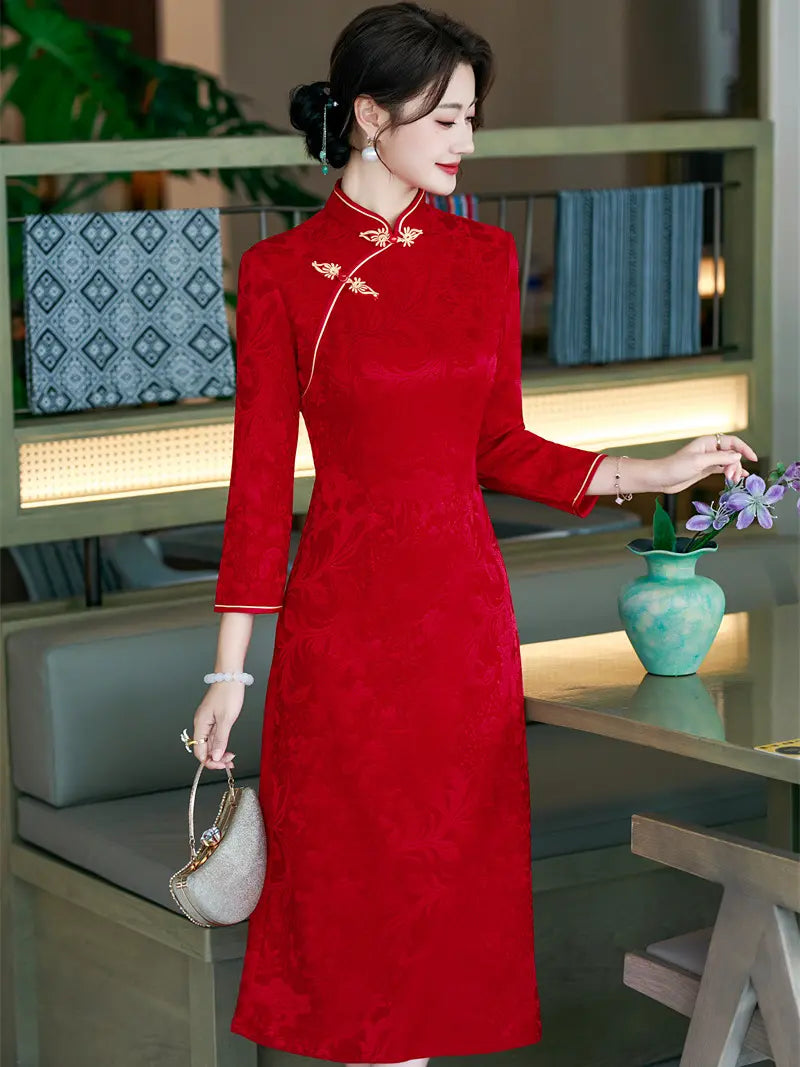 A woman wearing a long-sleeved red a line qipao cheongsam dress with gold dragon embellishments on the collar and chest, standing in a modern indoor setting with a table and vase.