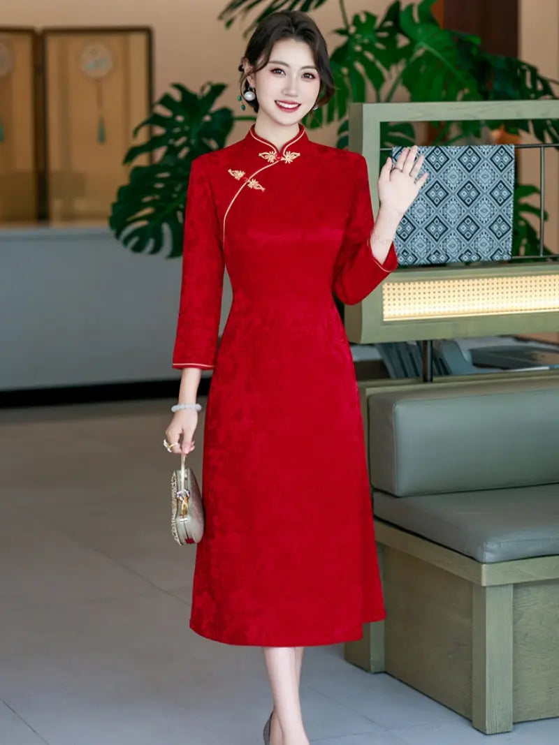 A woman wearing a red long-sleeve a line qipao cheongsam-style dress with gold embroidery on the collar and chest, standing in an indoor setting with green plants and furniture.
