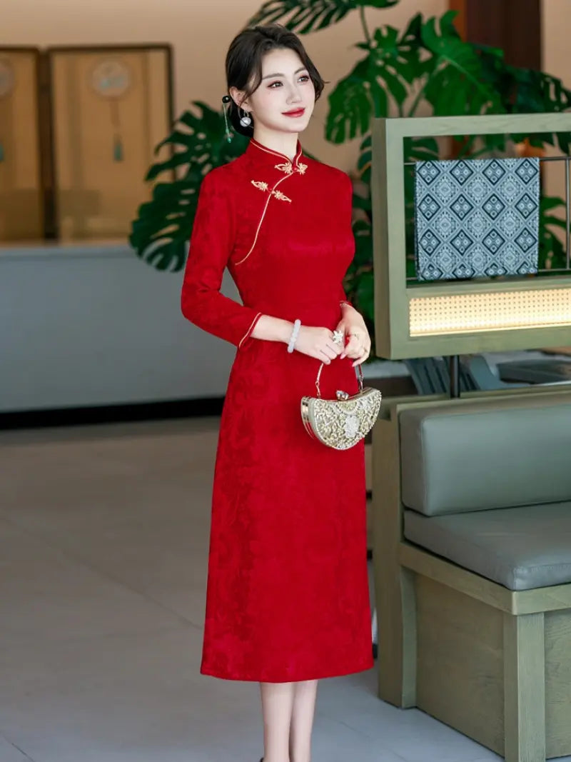 A woman wearing a red long-sleeve a line cheongsam-style dress with gold embroidery and a high collar, holding a small metallic handbag in an indoor setting with plants and furniture.