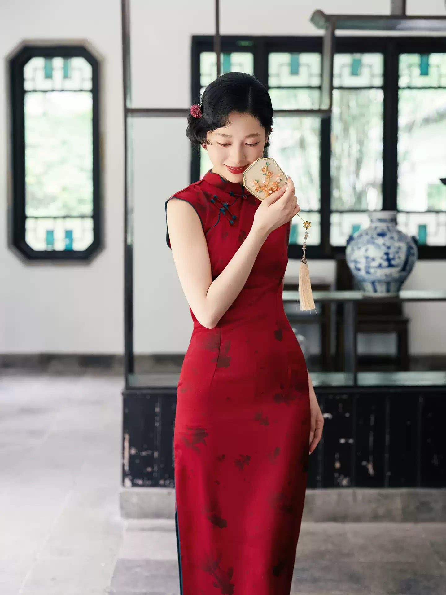 Woman in a Crimson Red Sleeveless Goldfish Qipao Dress holding a small object in a room with decorative elements.