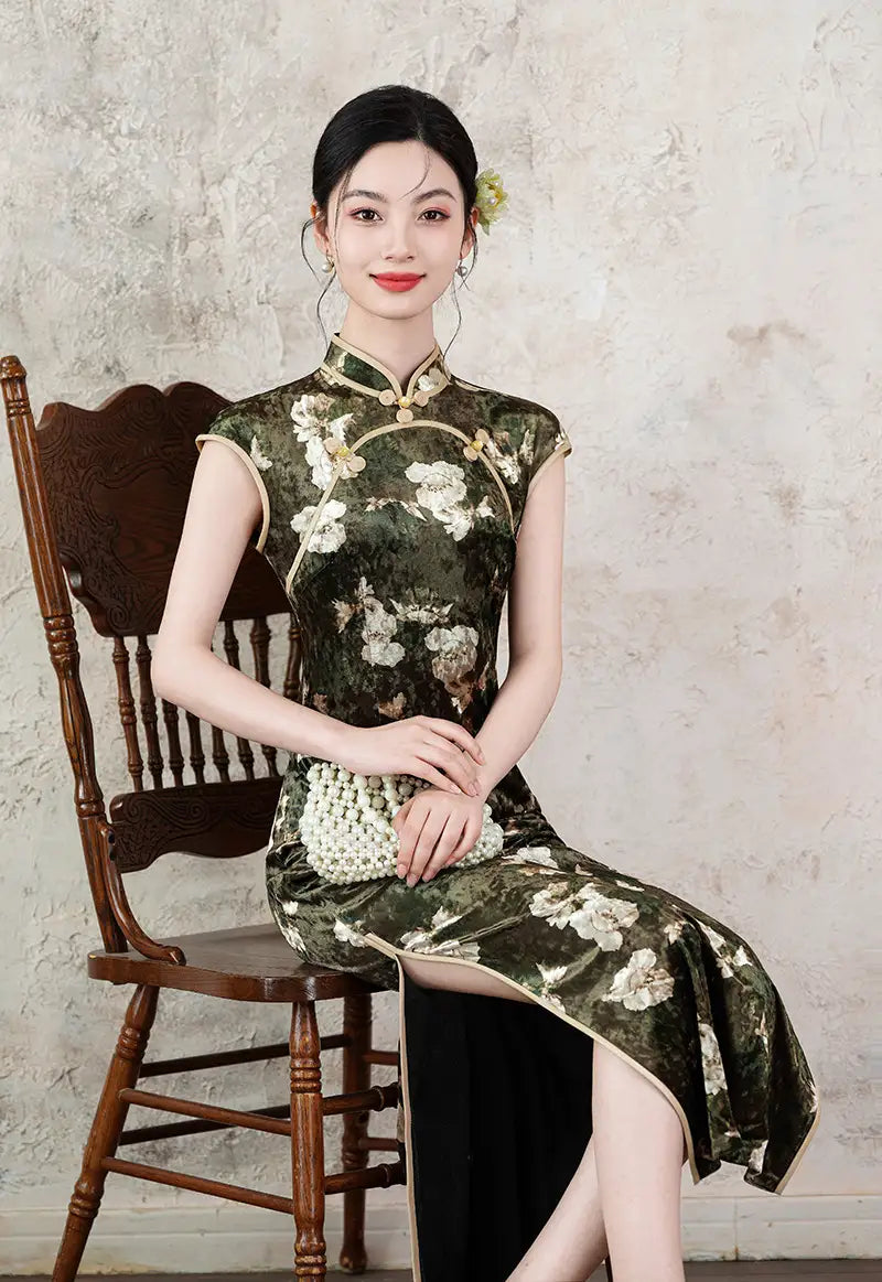 Woman in a Dark Green Foral Velvet Cap Sleeves Long Cheongsam Dress  sitting on a wooden chair against a textured wall.