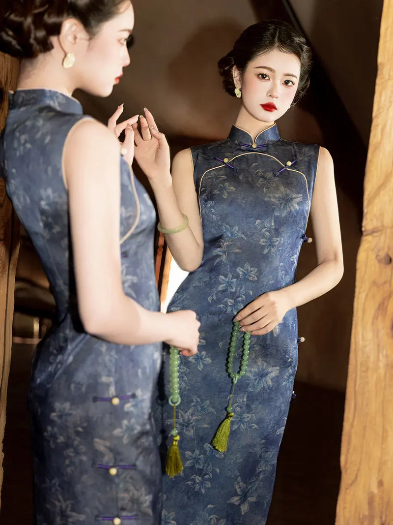 Two women wearing matching sleeveless blue floral cheongsam/qipao dresses pose indoors, with one facing the mirror and the other shown from the side. The setting includes warm wooden paneling and dim ambient lighting.