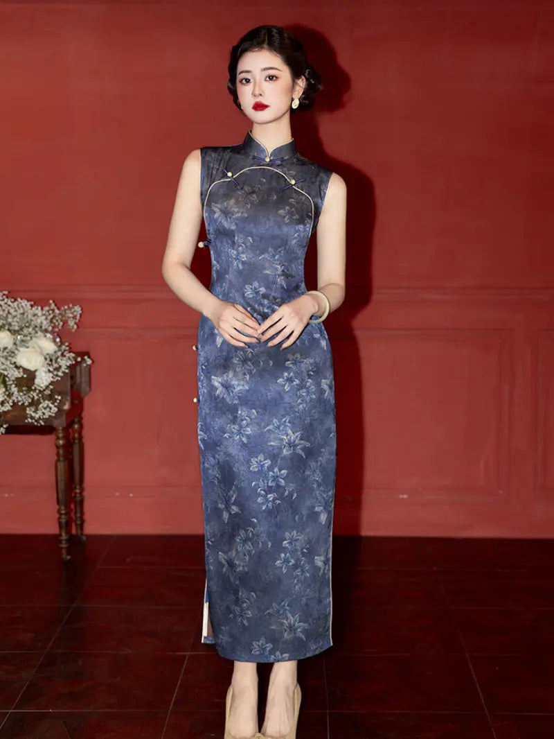 A woman wearing a sleeveless, floor-length blue floral cheongsam dress, standing against a dark red wall.