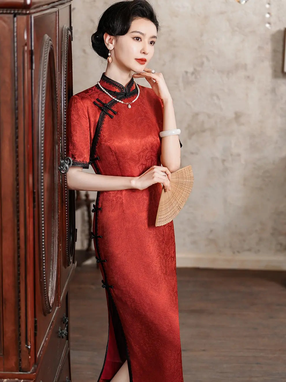 Full-length shot of a woman in a deep crimson silk jacquard qipao with black lace trim and frog buttons, standing in a vintage-style room
