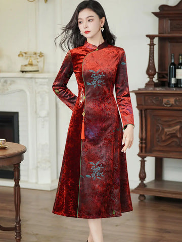 A woman modeling a long-sleeved red and burgundy brocade cheongsam-style dress with blue floral embroidery, standing in a classic indoor room with a fireplace and wooden furniture.