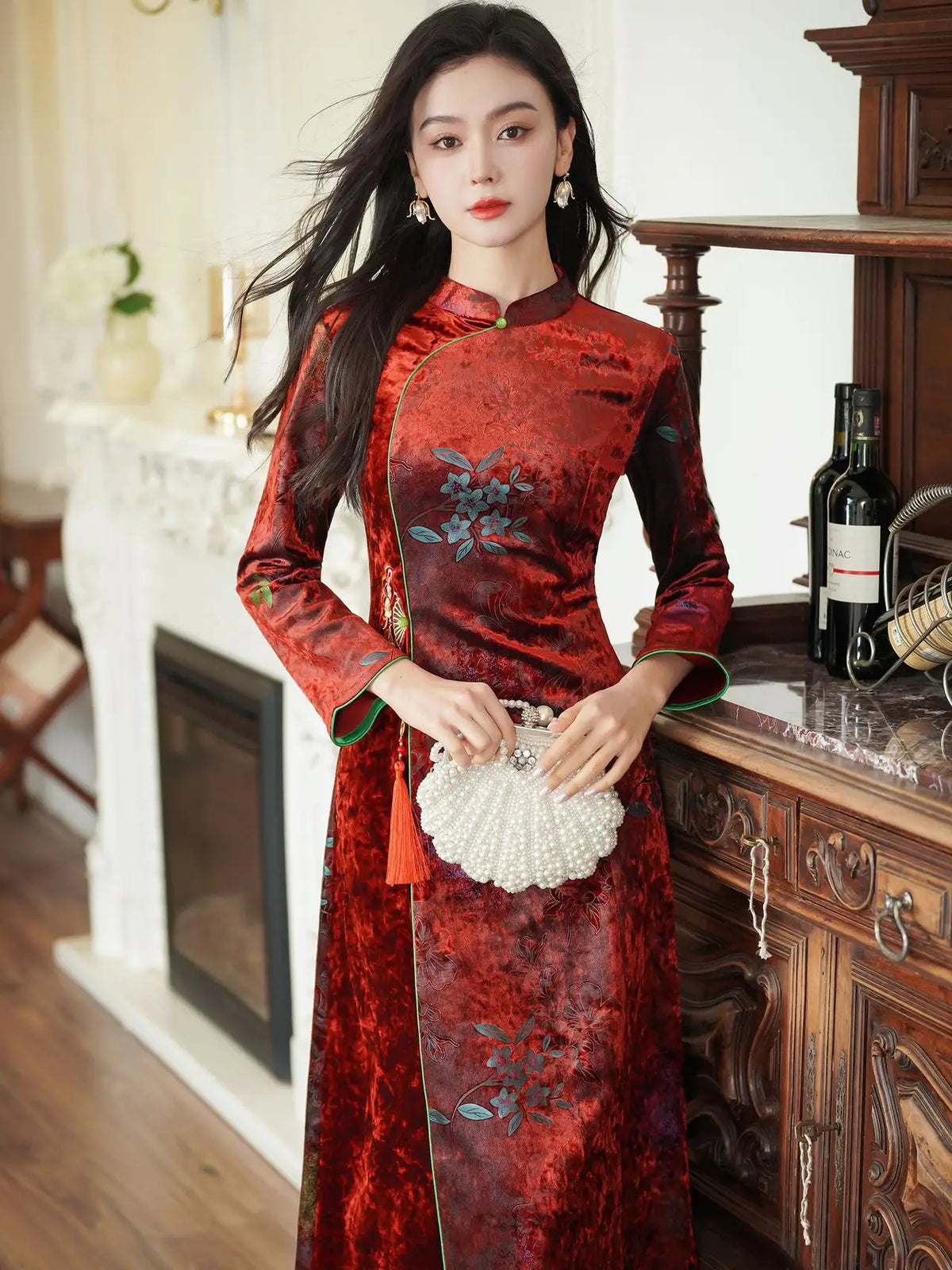 A woman wearing a long-sleeved red and long sleeve a line qipao cheongsam dress with floral embroidery, holding a white pearl clutch in an elegant indoor setting.