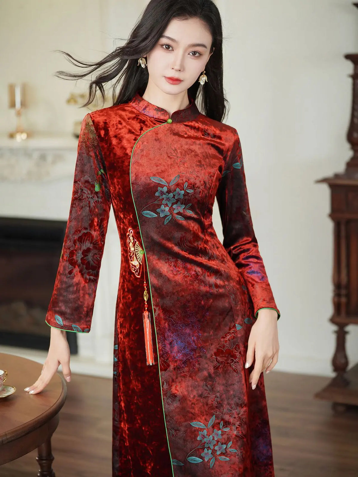 A woman wearing a long-sleeved red velvet cheongsam dress with blue floral embroidery and gold trim, standing in a warm, elegant indoor setting.