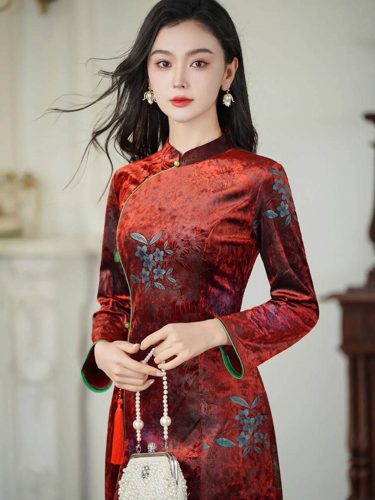 A woman wearing a red and dark blue patterned Chinese cheongsam dress with green trim, holding a pearl handbag in an indoor setting.