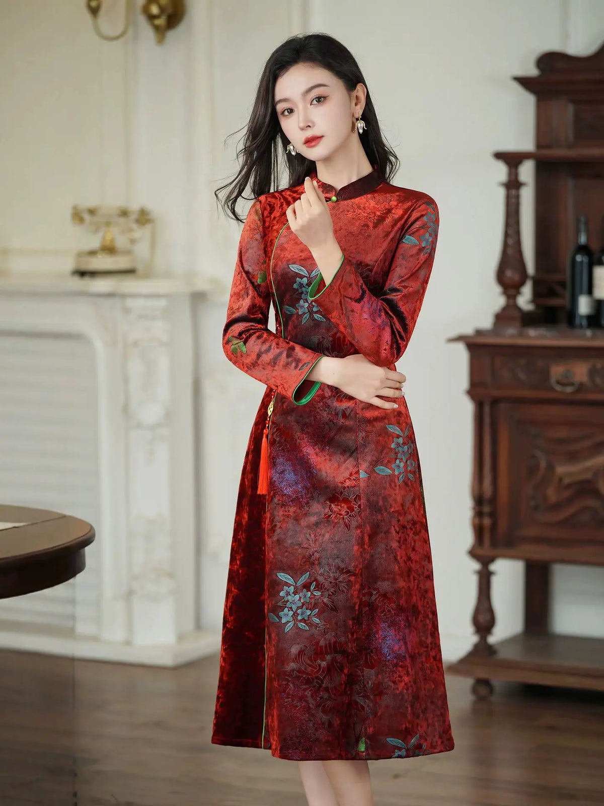 A woman wearing a red long-sleeve cheongsam-style dress with floral embroidery, standing in an elegant indoor room with a fireplace and wooden cabinet in the background.