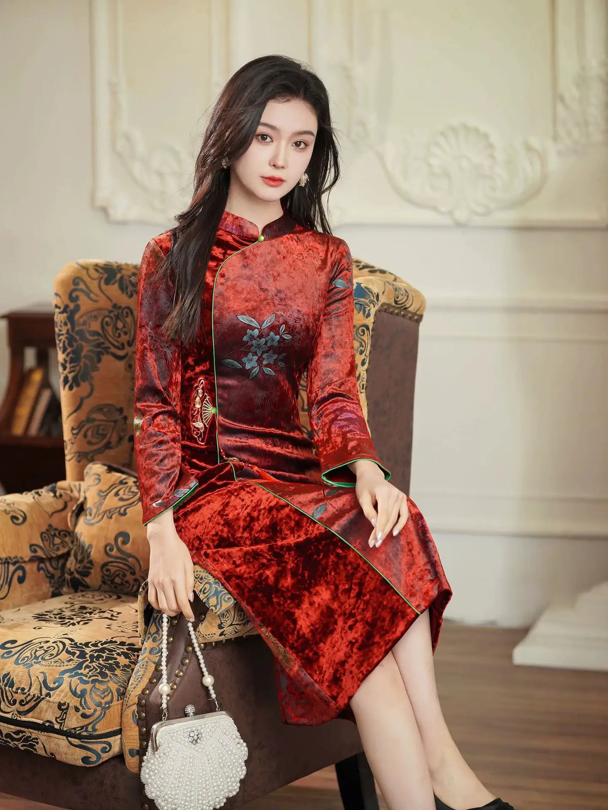 A woman sits in an ornate armchair wearing a red velvet Chinese cheongsam dress with floral embroidery, holding a small white pearl handbag in a classic indoor setting.