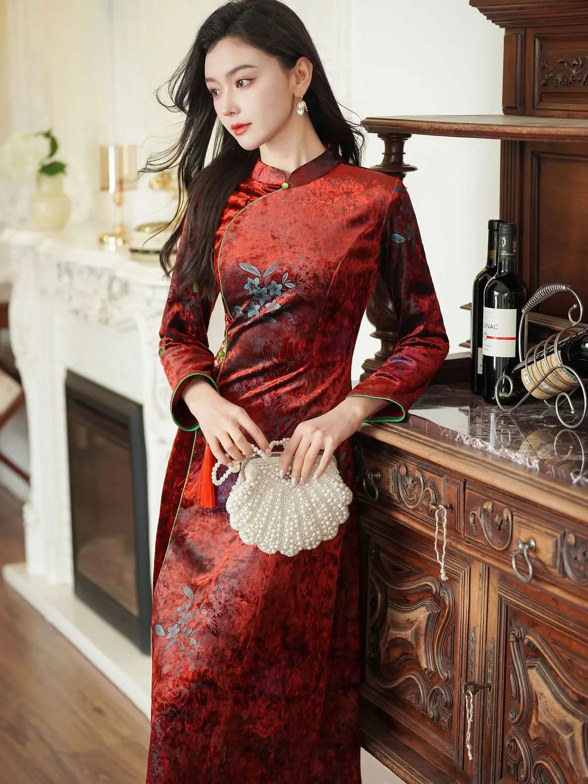 A woman wearing a red and black patterned Chinese cheongsam dress, holding a pearl shell clutch purse in a warm, vintage-style room with a fireplace and wooden cabinet.