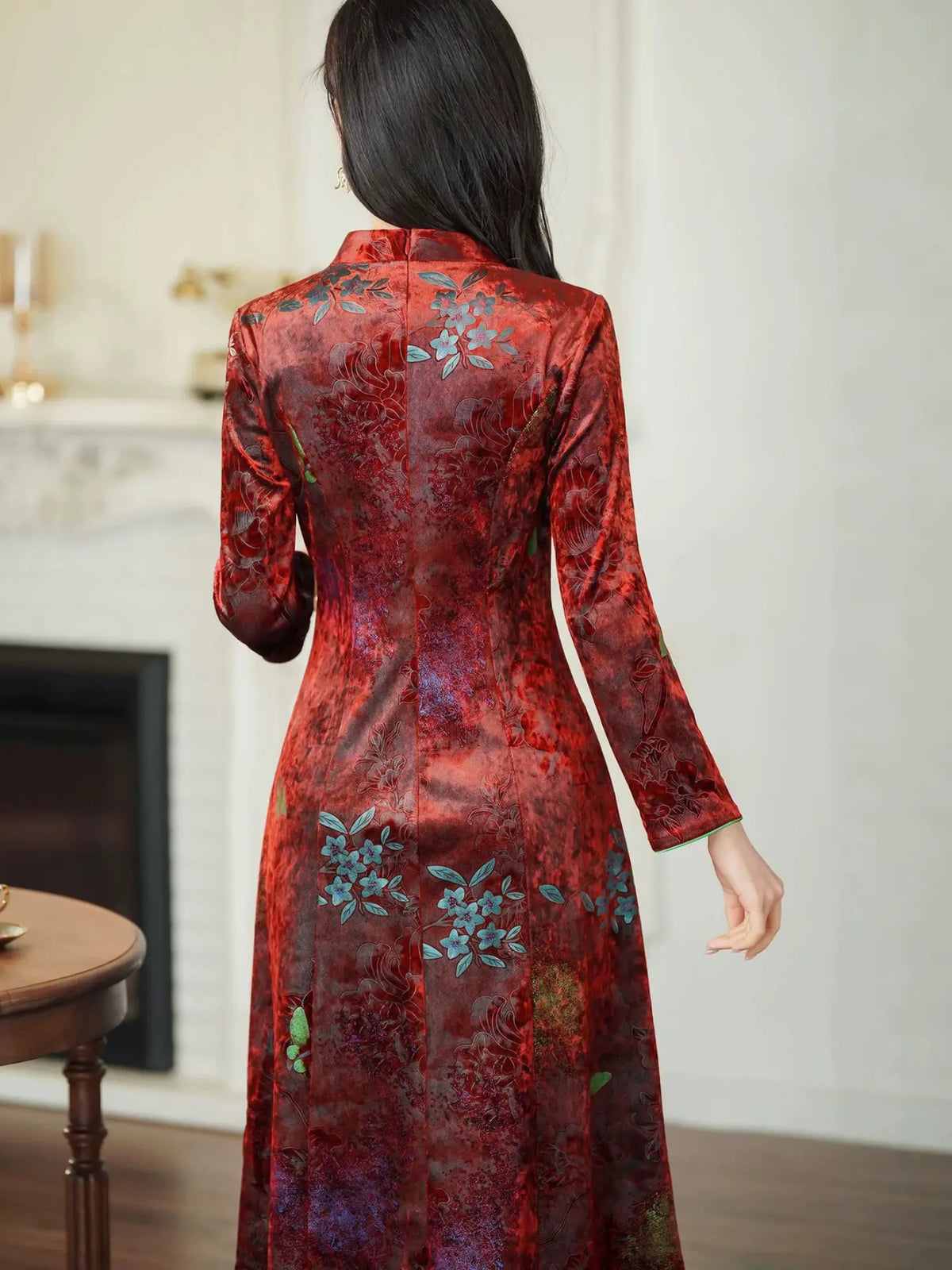 A woman viewed from behind wearing a long-sleeved red Vietnamese ao dai dress with a floral pattern and blue accents, standing in an indoor setting.