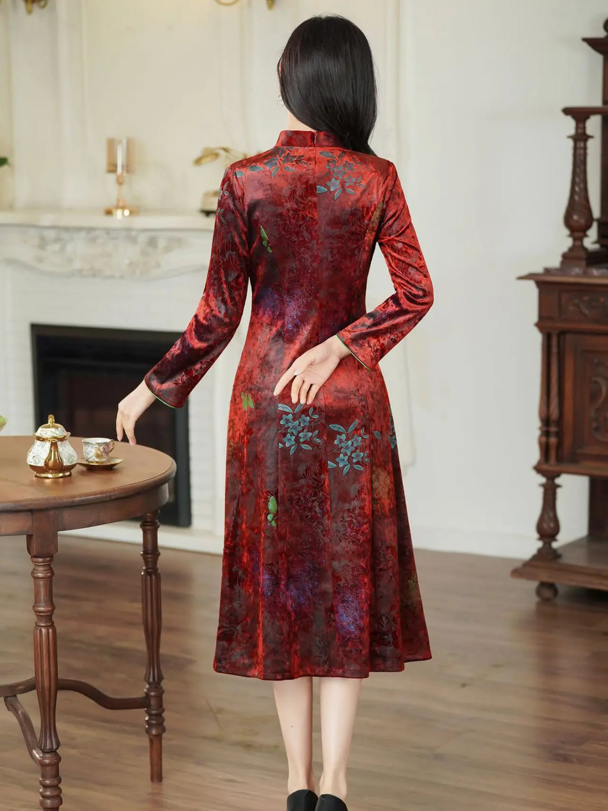 A woman wearing a long-sleeved red cheongsam dress with a blue and green floral pattern, photographed from behind in a classic indoor room setting.