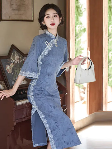 A woman models a blue traditional Chinese cheongsam dress with white lace trim, holding a light gray handbag in a sunlit indoor setting.