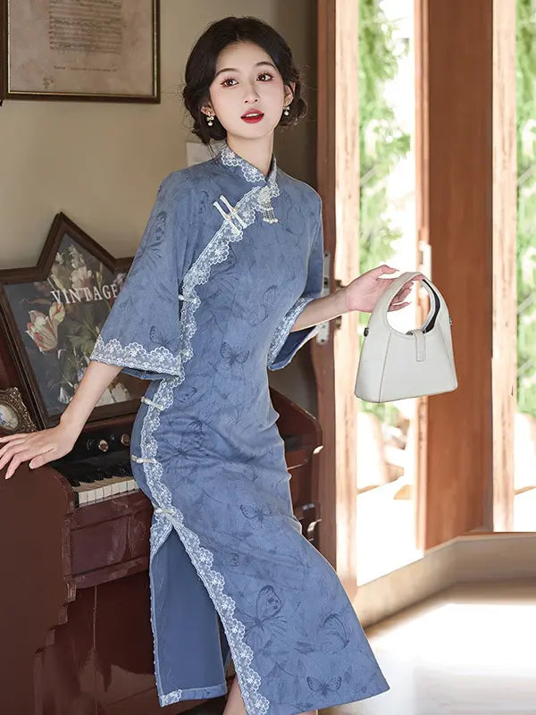A woman models a blue traditional Chinese cheongsam dress with white lace trim, holding a light gray handbag in a sunlit indoor setting.
