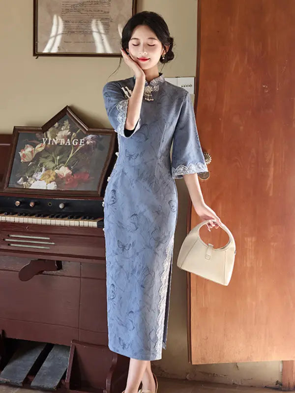A woman models a light blue floral-patterned long sleeve cheongsam dress in a vintage room setting, holding a cream-colored handbag next to a wooden door and an antique piano.