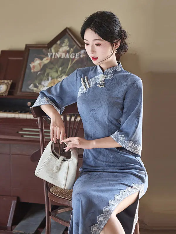 A woman seated on a wooden chair wearing a blue long sleeve cheongsam-style dress with lace trim and holding a light beige handbag in a vintage indoor setting