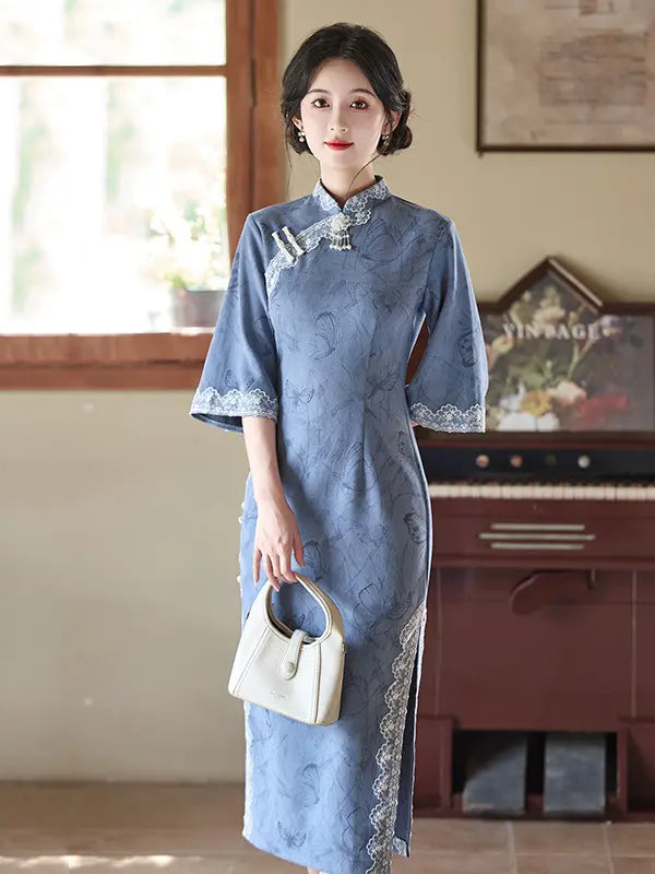 A woman wearing a light blue traditional long sleeve Chinese cheongsam dress with lace trim, holding a small cream-colored handbag in an indoor setting with a piano in the background.