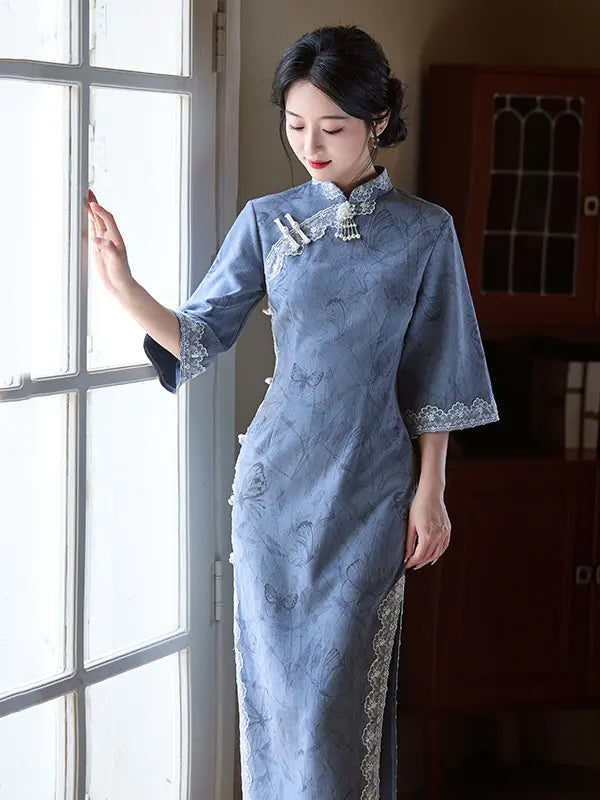 A woman wearing a light blue-gray long sleeve cheongsam (qipao) dress stands by a window in a dimly lit room. The dress features delicate floral embroidery, silver-tone trim, and a traditional collar with decorative cord details.