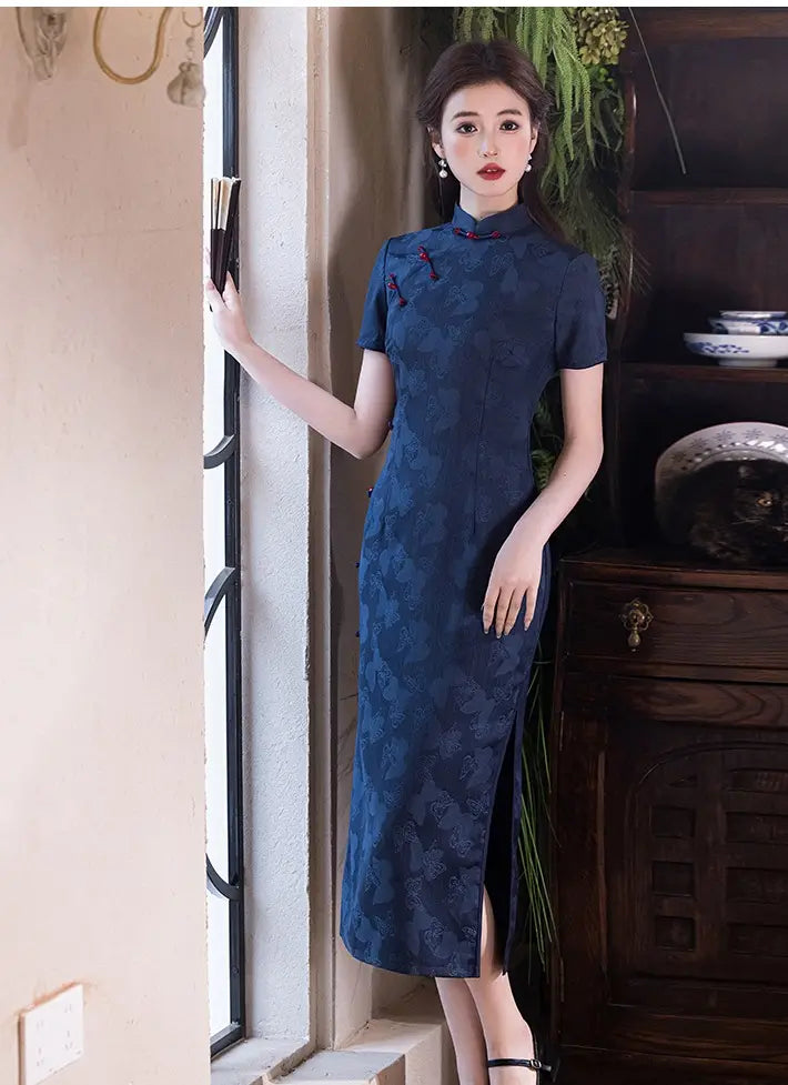 Woman in a Elegant Blue Jacquard Butterfly Cheongsam – Mid-Length Modern Qipao standing in a room with a wooden cabinet and decorative items.