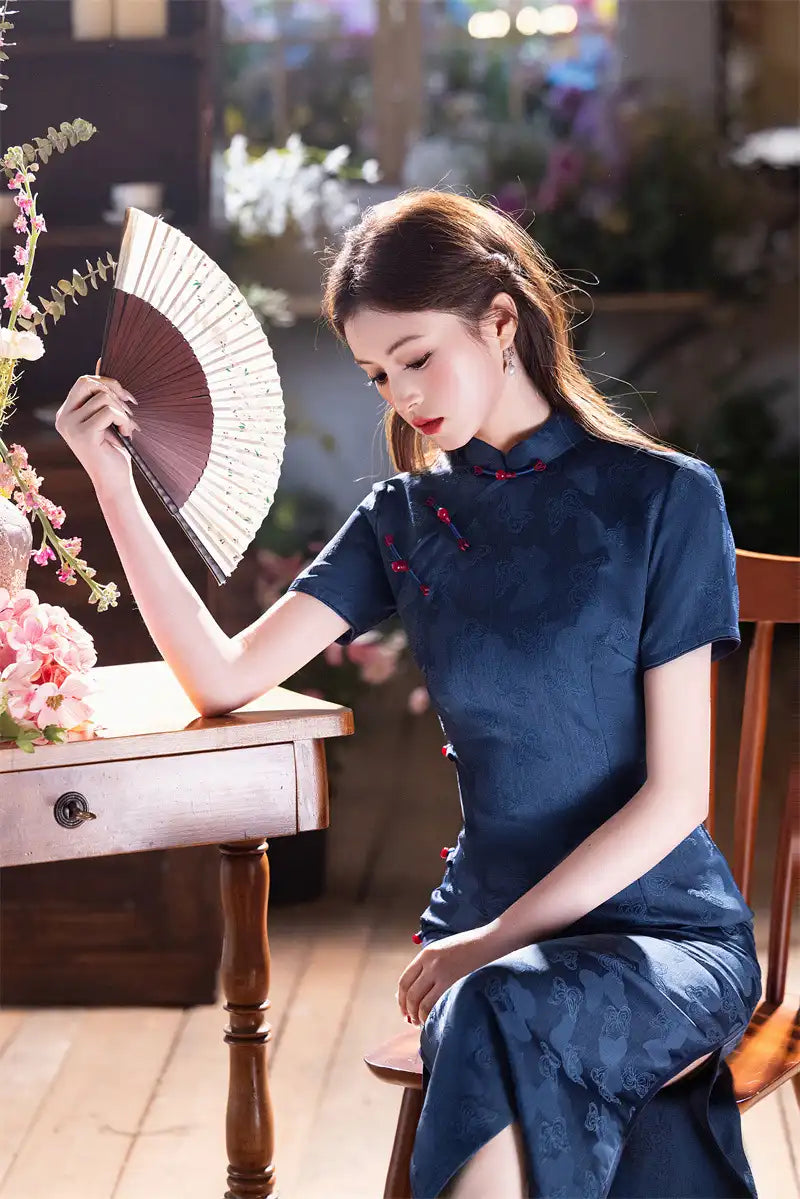 Woman in a Elegant Blue Jacquard Butterfly Cheongsam – Mid-Length Modern Qipao holding a fan in a softly blurred indoor setting