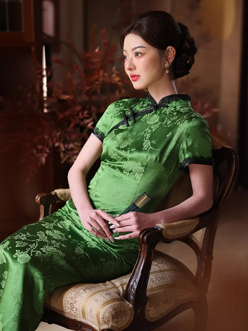 Woman in aEmerald Forest Green Silk-Blend Qipao with Lace Trim  traditional dress sitting on a wooden chair in a warm-toned room.
