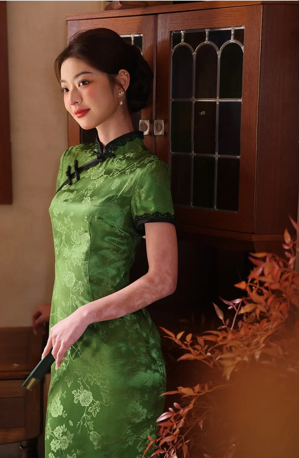 Woman in a Emerald Forest Green Silk-Blend Qipao with Lace Trim traditional dress standing indoors with a wooden cabinet in the background.