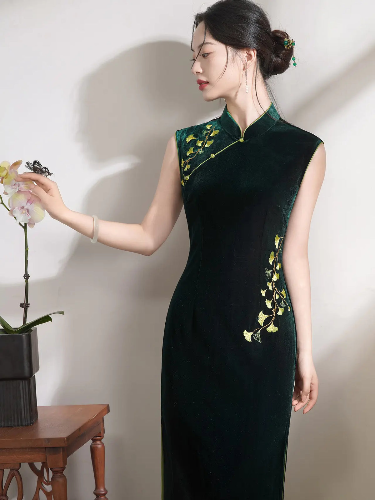 A woman wearing a dark green velvet cheongsam dress with yellow floral embroidery, posing next to a small wooden table with a potted orchid against a neutral wall.