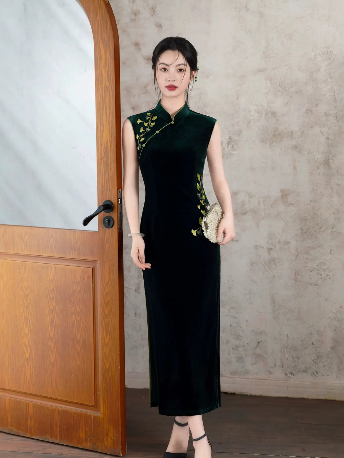 A woman stands indoors wearing a sleeveless black velvet cheongsam-style dress with green floral embroidery near the neckline and side. She is posed beside a wooden door against a textured light gray wall, holding a small metallic clutch.