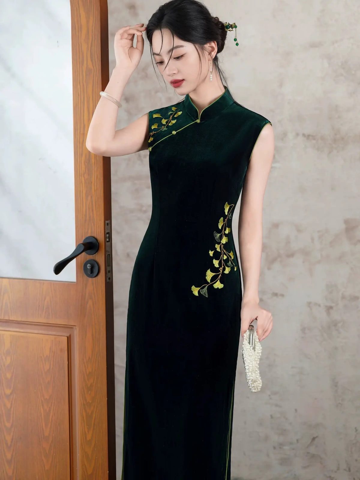 A woman wearing a dark green velvet cheongsam dress with yellow floral embroidery on the shoulders and side, standing next to a wooden door.