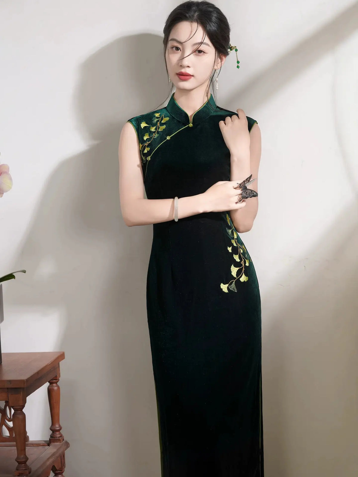 A woman wearing a sleeveless dark green cheongsam-style dress with gold floral embroidery near the shoulders and hips, standing indoors against a light wall.