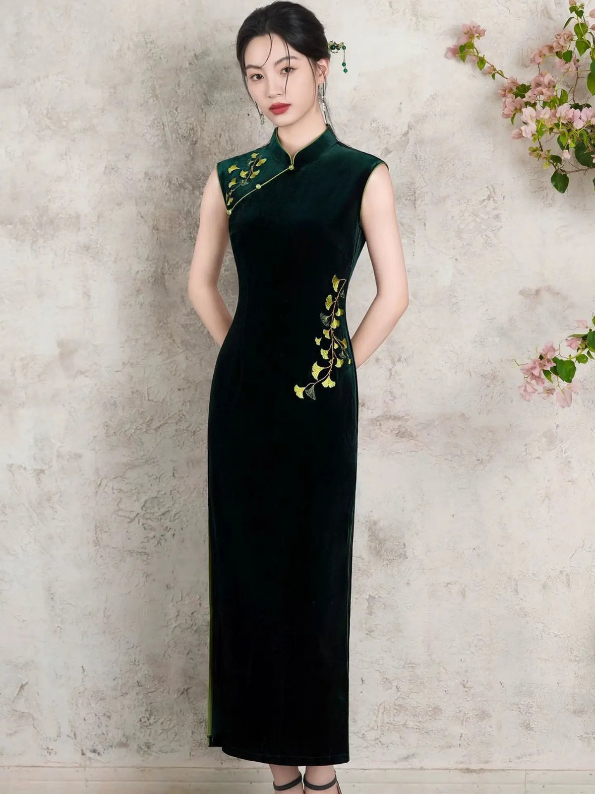 A woman wearing a sleeveless dark green velvet cheongsam dress with gold floral embroidery, standing against a textured light beige wall with pink flowers in the corner.