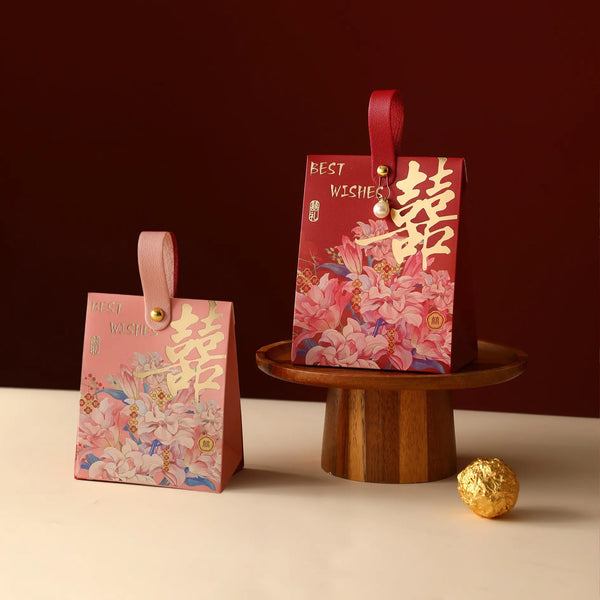 Two Floral Double Happiness Chinese Wedding Favor Gift Boxes  with floral prints and Chinese characters are displayed on a tabletop, one standing on the floor and one elevated on a wooden pedestal. A small gold ball sits nearby against a dark red background.