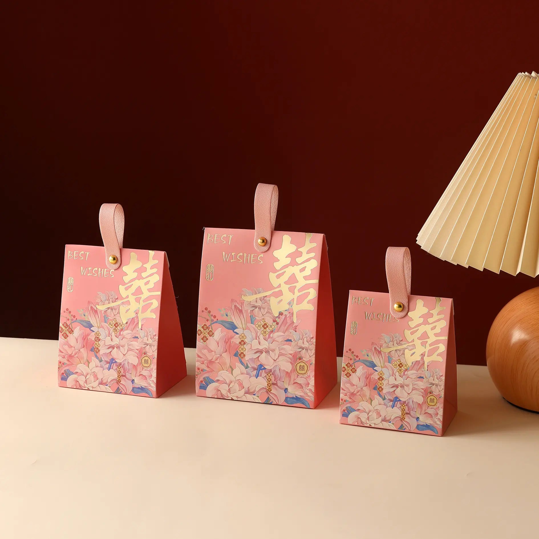 Three pink Floral Double Happiness Chinese Wedding Favor Gift Boxes  with gold Chinese characters and small handles, arranged on a light surface against a dark red background, with part of a cream pleated lamp visible on the right.