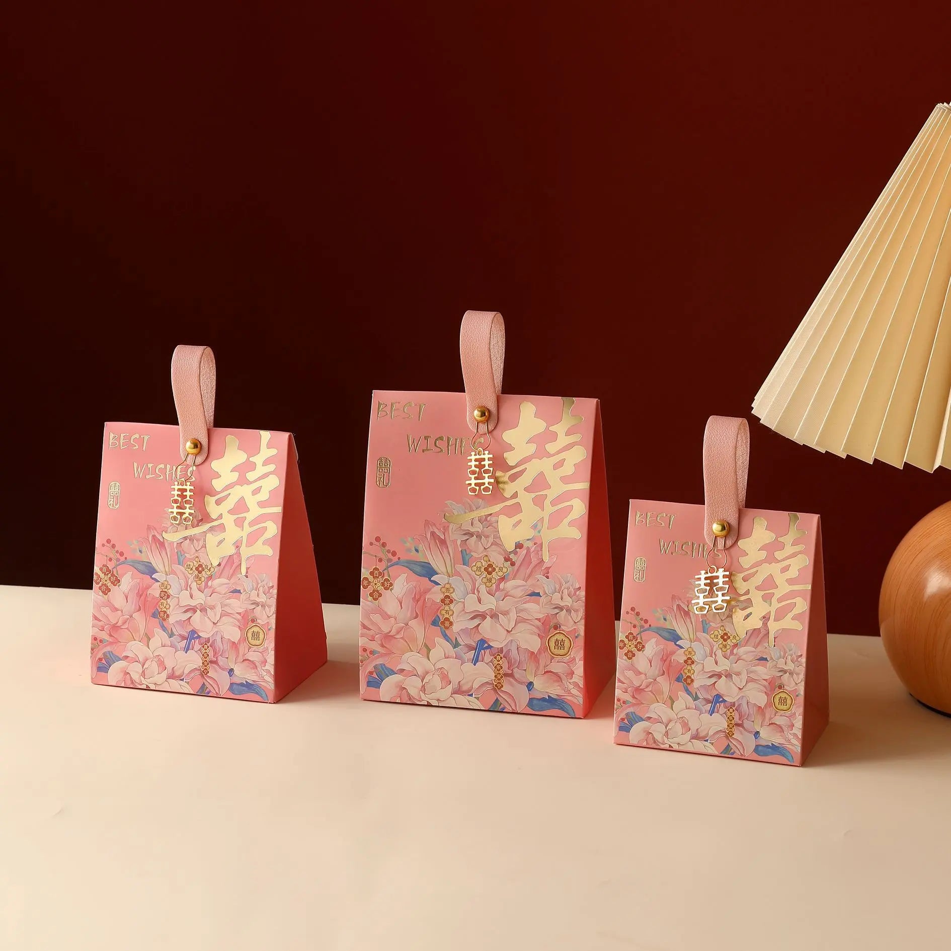 Three pink Floral Double Happiness Chinese Wedding Favor Gift Boxes  with pink handles and gold Chinese double-happiness symbols, arranged on a light beige surface against a dark red background with a cream pleated lamp visible on the right.
