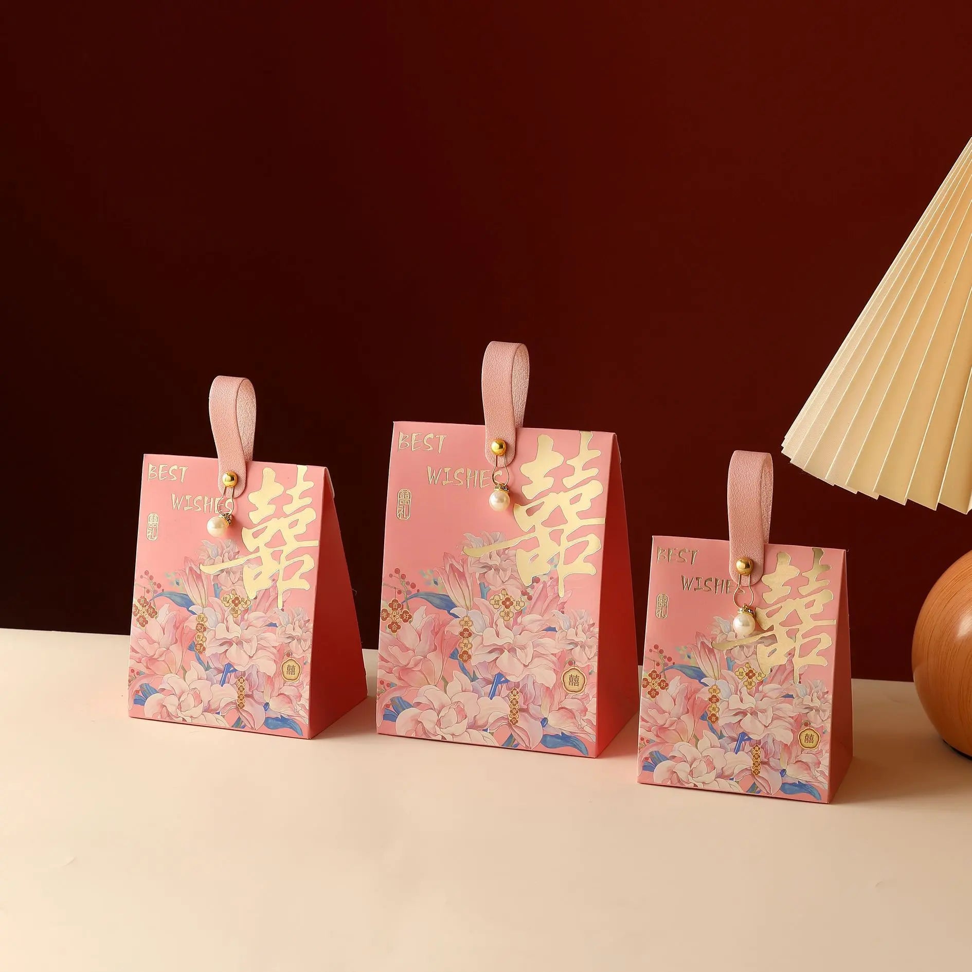 Three pink Floral Double Happiness Chinese Wedding Favor Gift Boxes  with gold Chinese characters and ribbon handles are arranged on a cream surface against a dark red background, with a folded paper fan and a wooden object partially visible on the right.