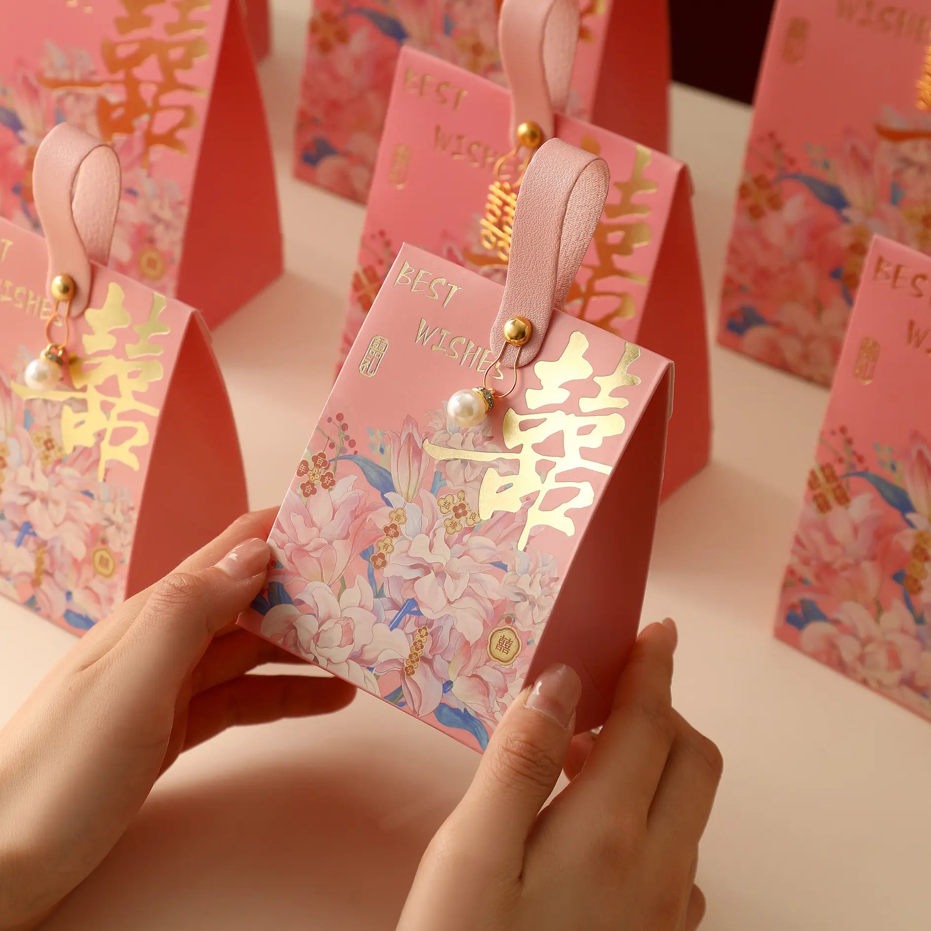 A person holds a pink Floral Double Happiness Chinese Wedding Favor Gift Boxes with gold Chinese characters and a pink ribbon loop, with similar bags arranged in the background.