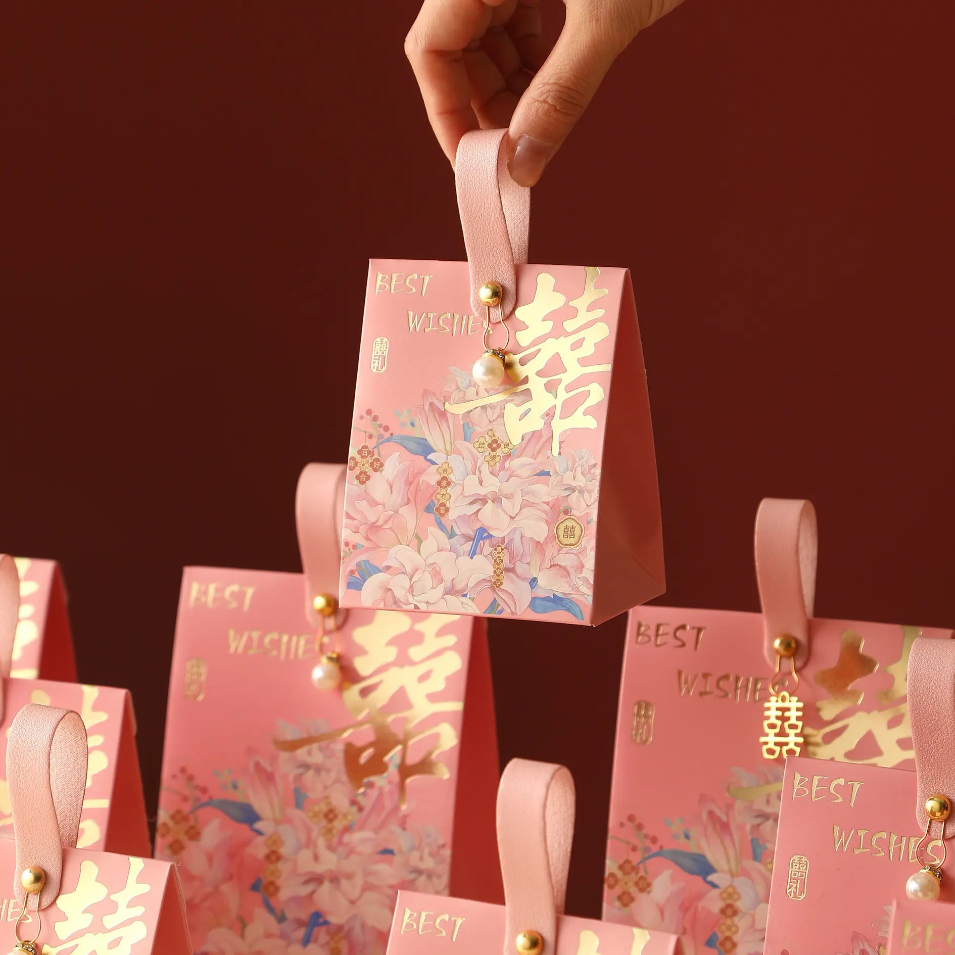 A hand holds a pink Floral Double Happiness Chinese Wedding Favor Gift Boxes  with a ribbon handle. The bag has gold Chinese characters, floral illustrations, and the words 'BEST WISHES' on a dark red background.
