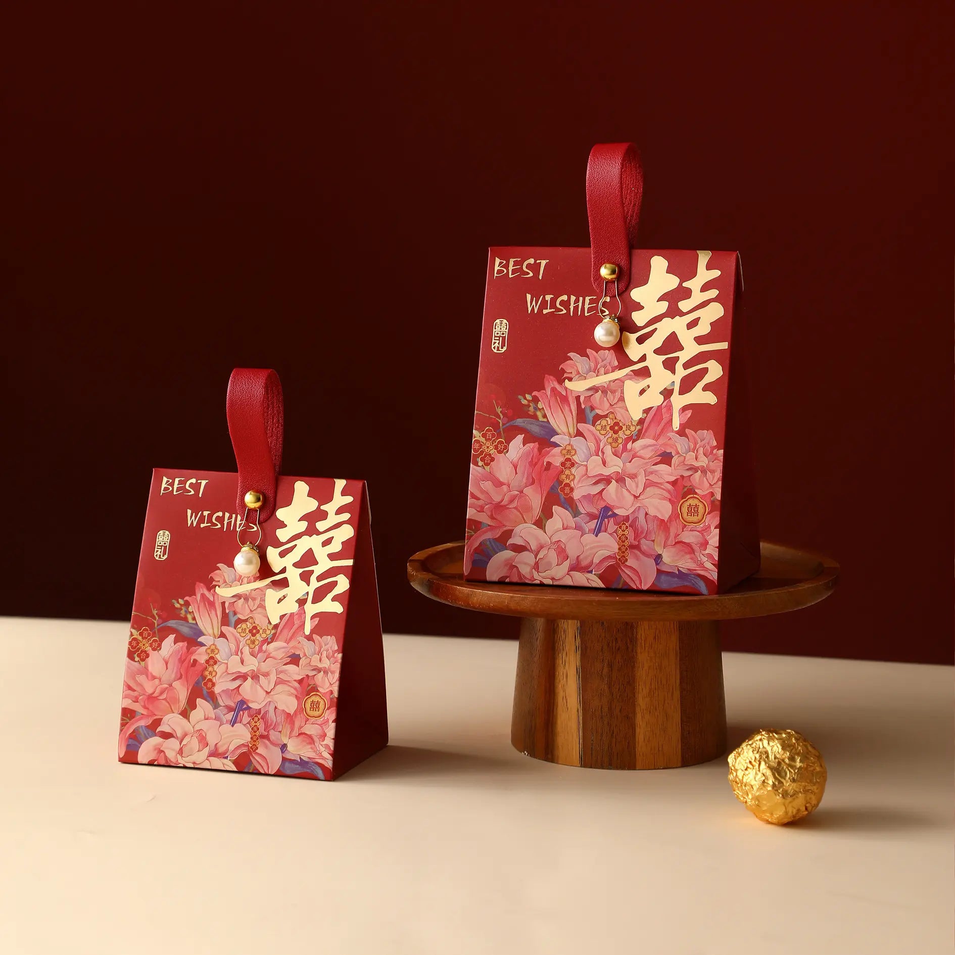 Two red fFloral Double Happiness Chinese Wedding Favor Gift Boxes  with handles are displayed on a tabletop, one standing on a wooden pedestal and one beside it. The boxes feature pink and peach floral artwork, gold Chinese characters, and the words 'BEST WISHES.