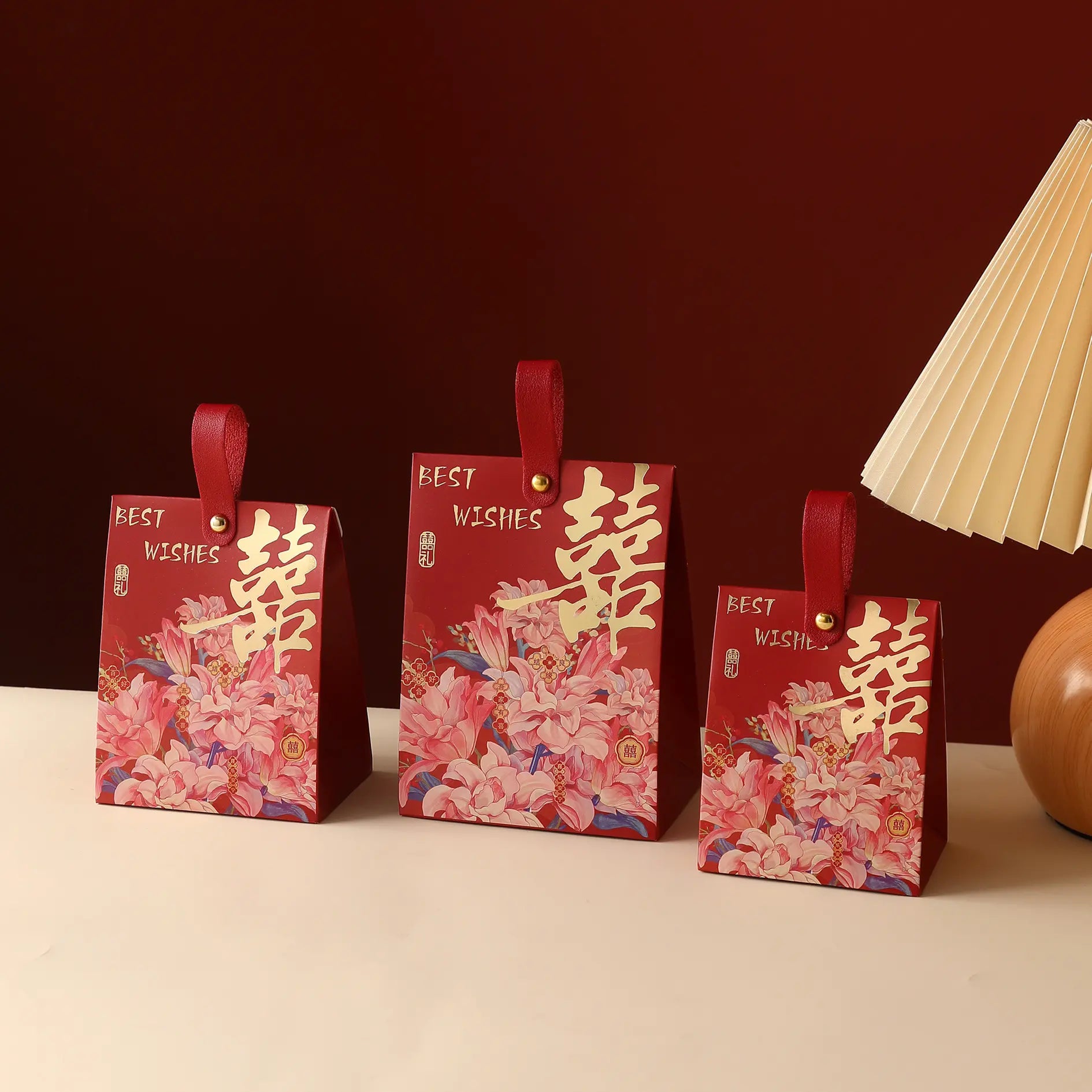 Three red Floral Double Happiness Chinese Wedding Favor Gift Boxes  with floral pink and gold patterns, Chinese double happiness symbols, and red handles, arranged on a light beige surface against a dark red background with a lamp partially visible on the right.