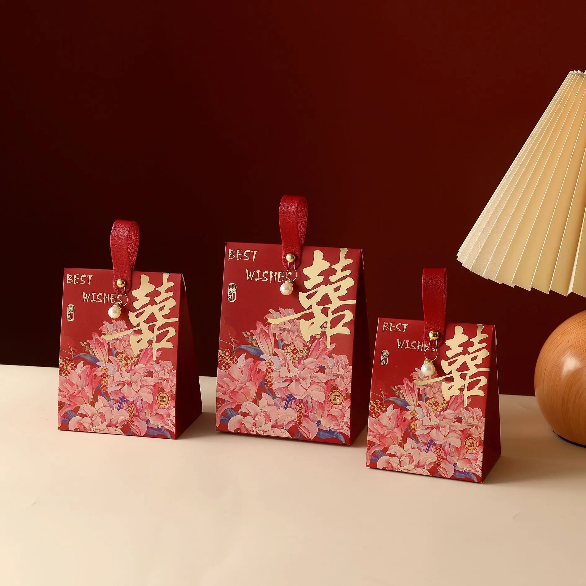 Three red Floral Double Happiness Chinese Wedding Favor Gift Boxes  with handles, decorated with pink floral artwork and large gold Chinese characters, arranged on a light surface against a dark red background with a cream pleated lamp visible on the right.