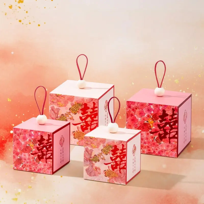 Four decorative red and pink gift boxes with floral Kio fish patterns and large Chinese double happiness characters, arranged on a soft peach background with gold speckles.